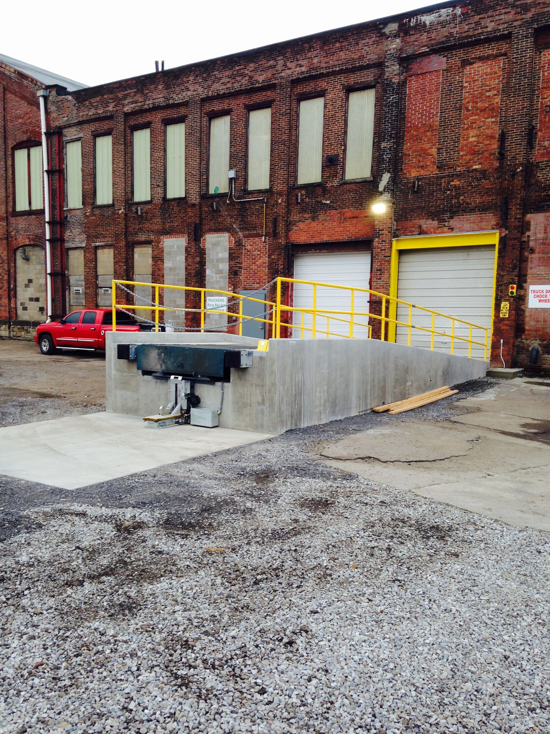 Loading dock ramp with yellow railing against a brick building, gravel in front. Red truck parked.