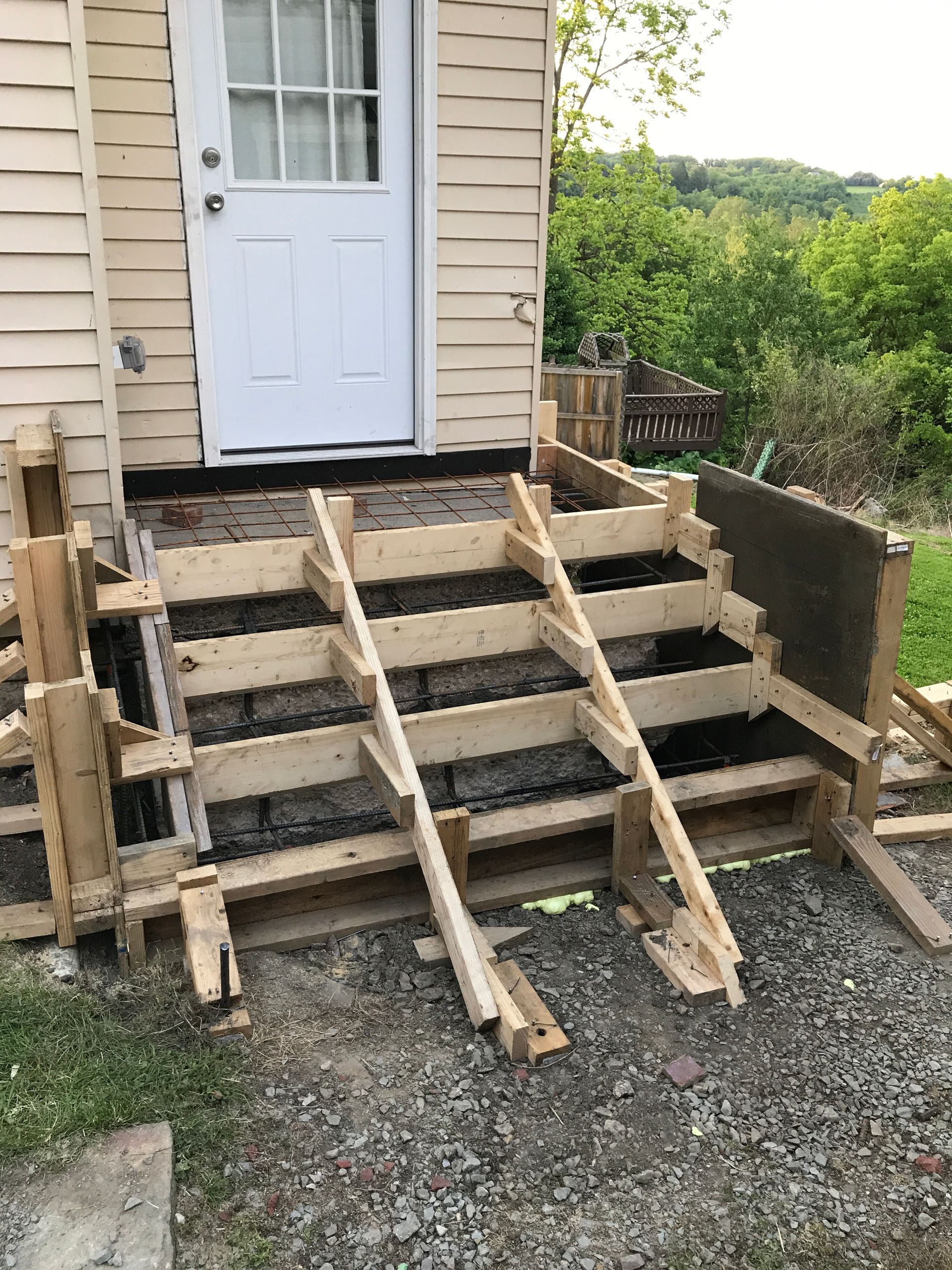Wooden formwork for concrete steps leading to a white door. Gravel ground. Green trees in the background.