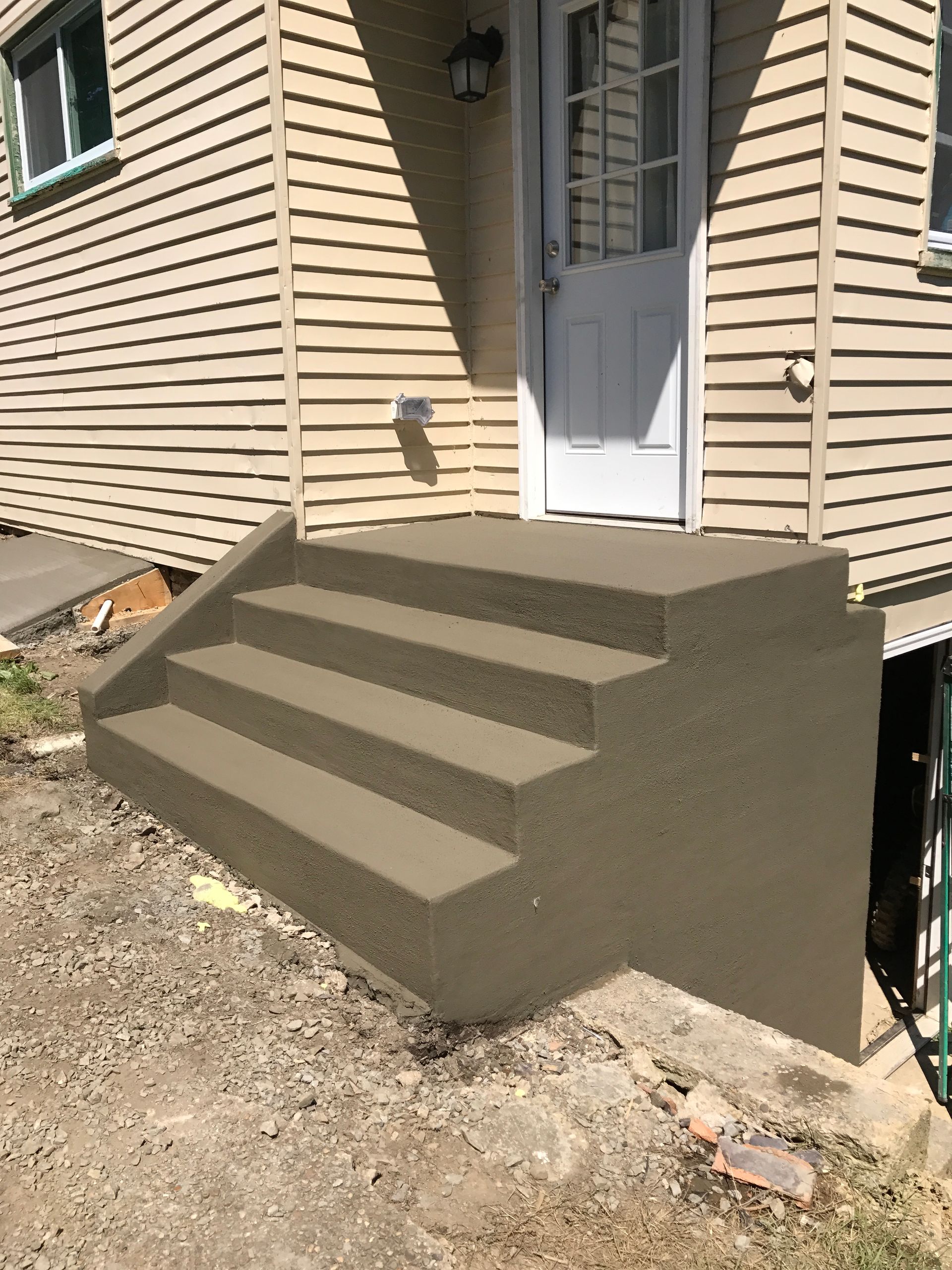 Concrete steps leading to a white door on a beige house. Exterior setting with dirt ground.