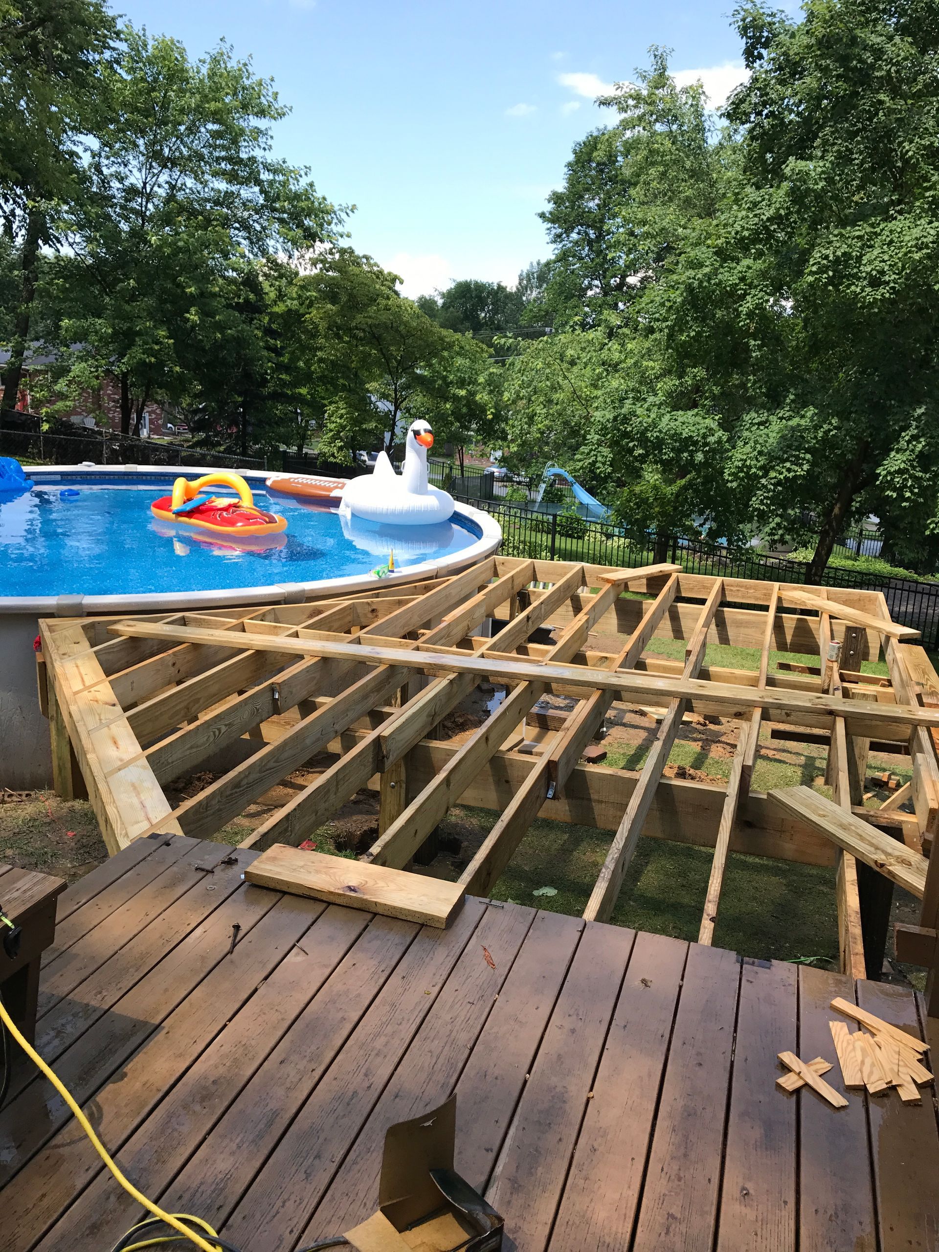 Deck under construction next to a pool, with lawn and trees in the background.