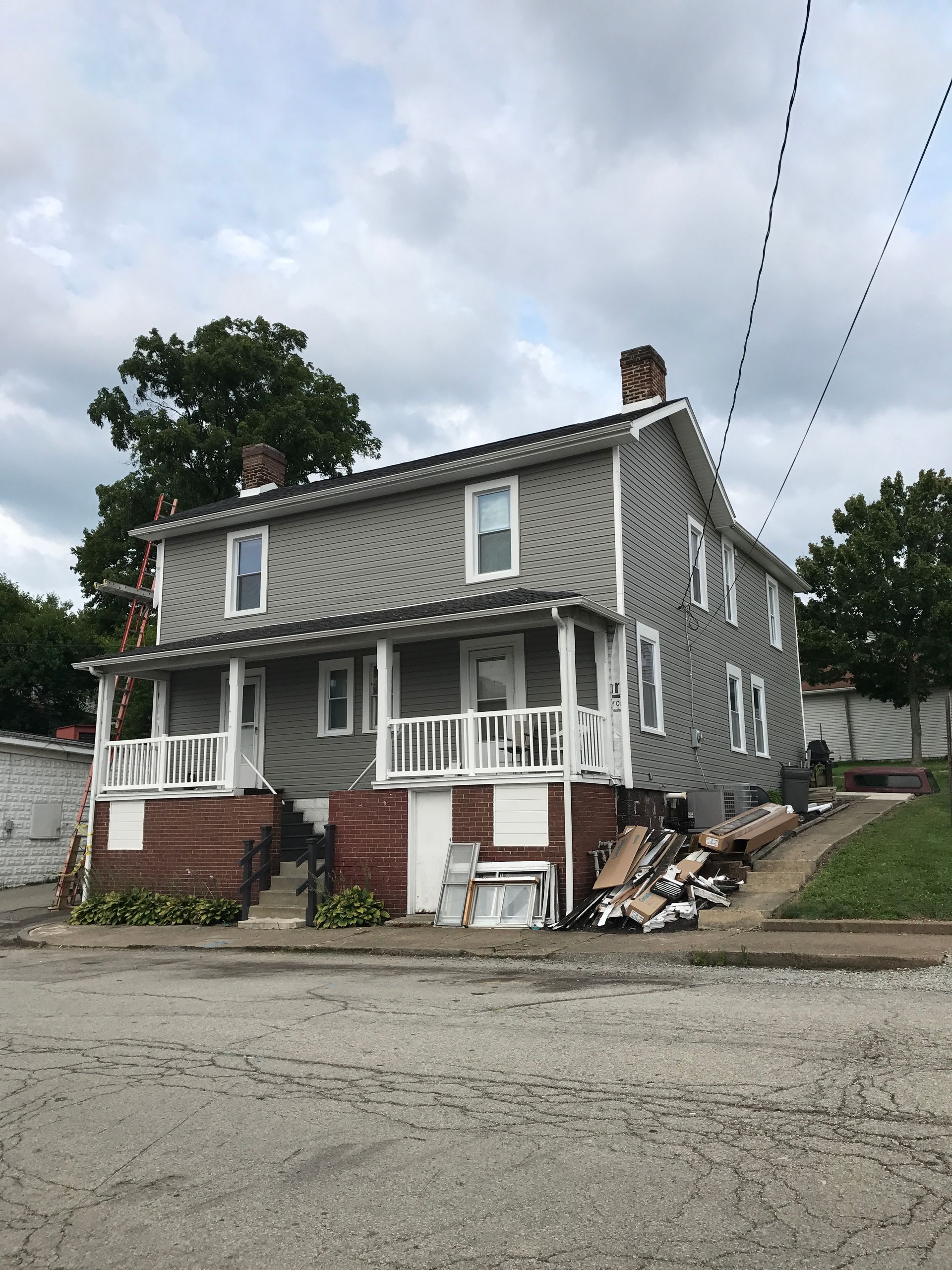Two-story gray duplex with white trim, red brick foundation, and a debris pile in front, under a cloudy sky.