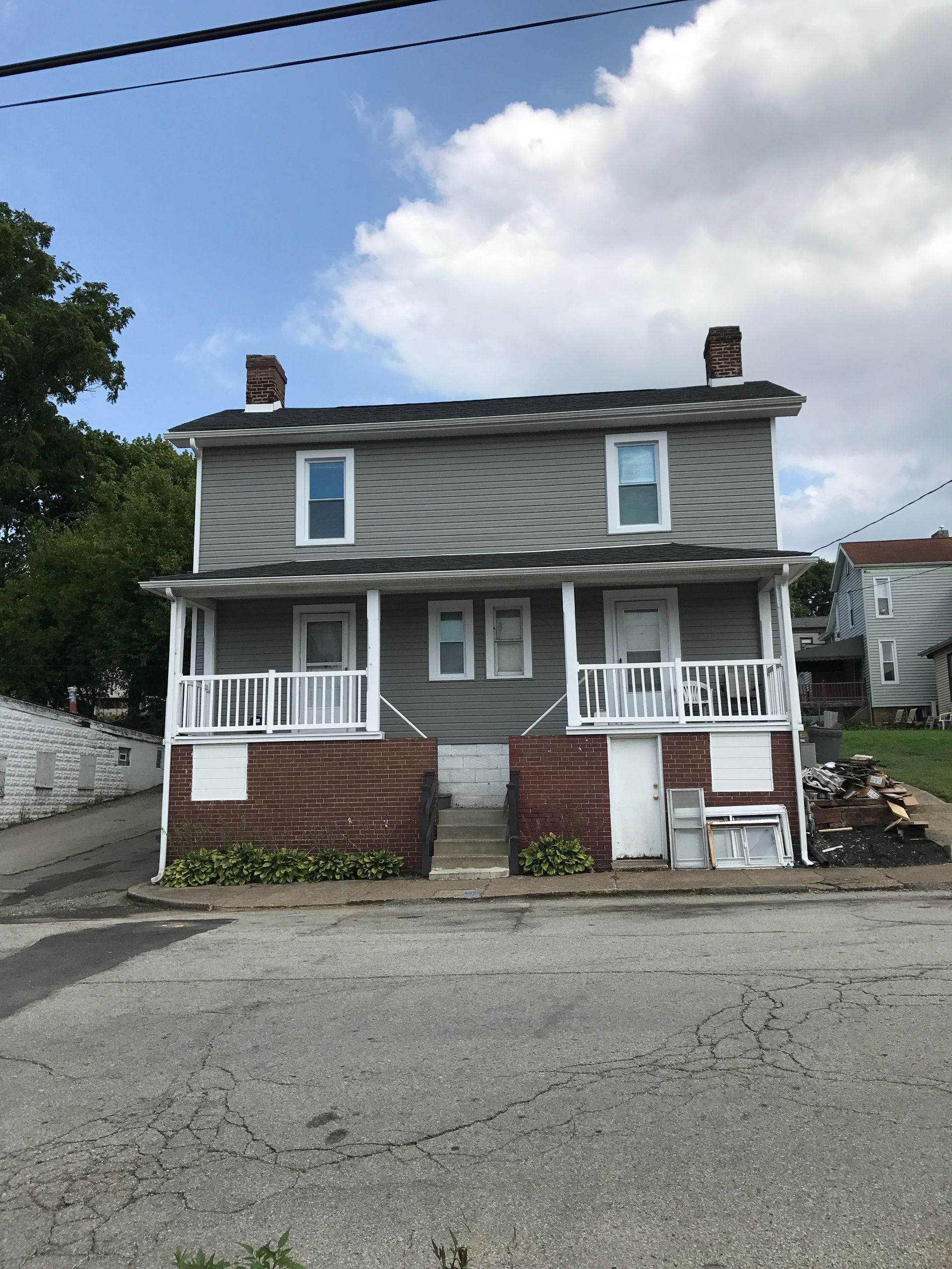 Two-story gray and brick duplex with front porches, set on a slight incline, against a cloudy sky.