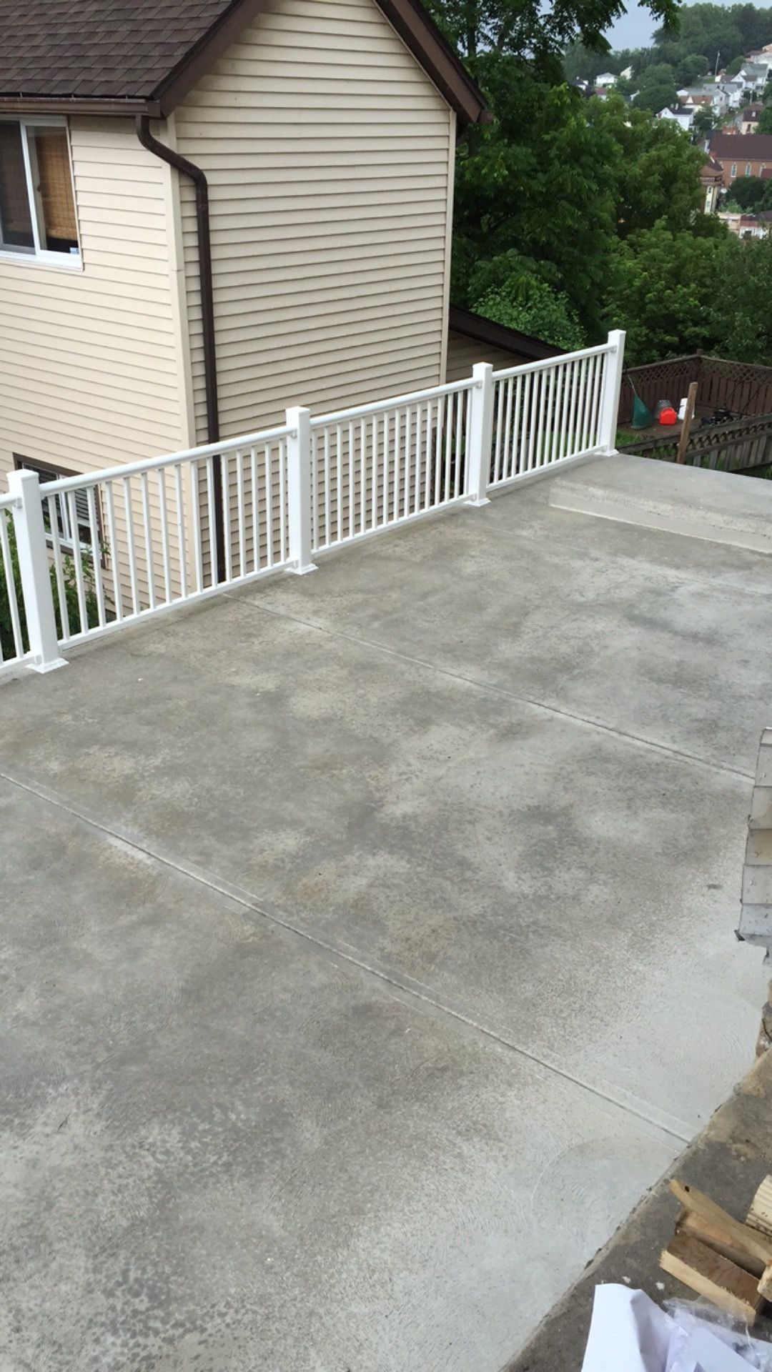 White railing surrounds a concrete patio next to a beige house. Trees in the background.