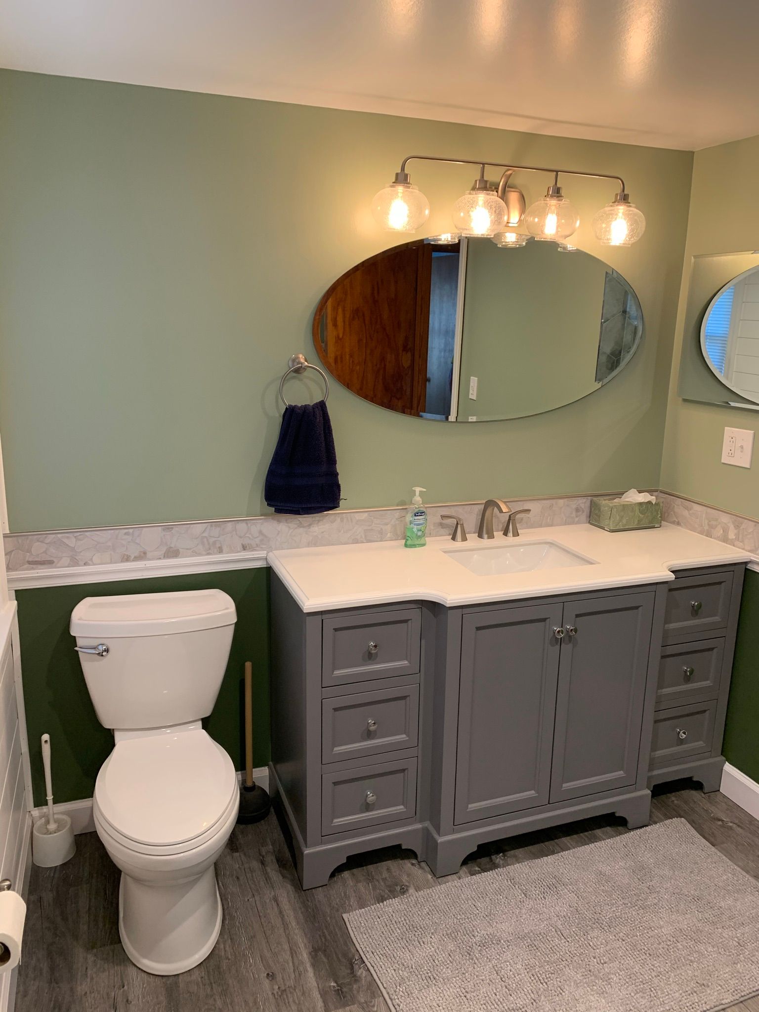 Bathroom with green walls, gray vanity, toilet, and oval mirror.