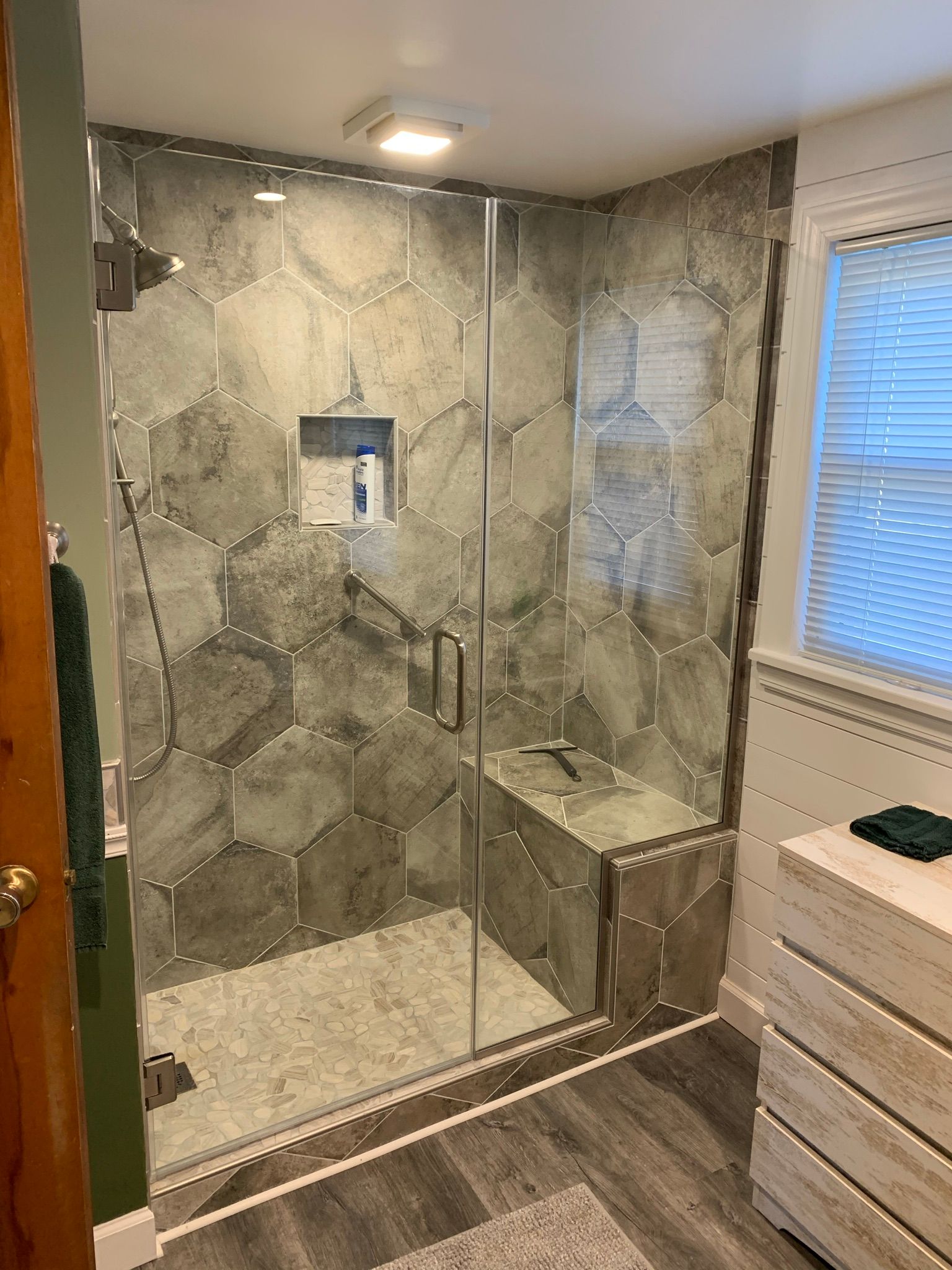 Modern bathroom with gray hexagonal tile shower, glass door, built-in seat, and wood-look flooring.