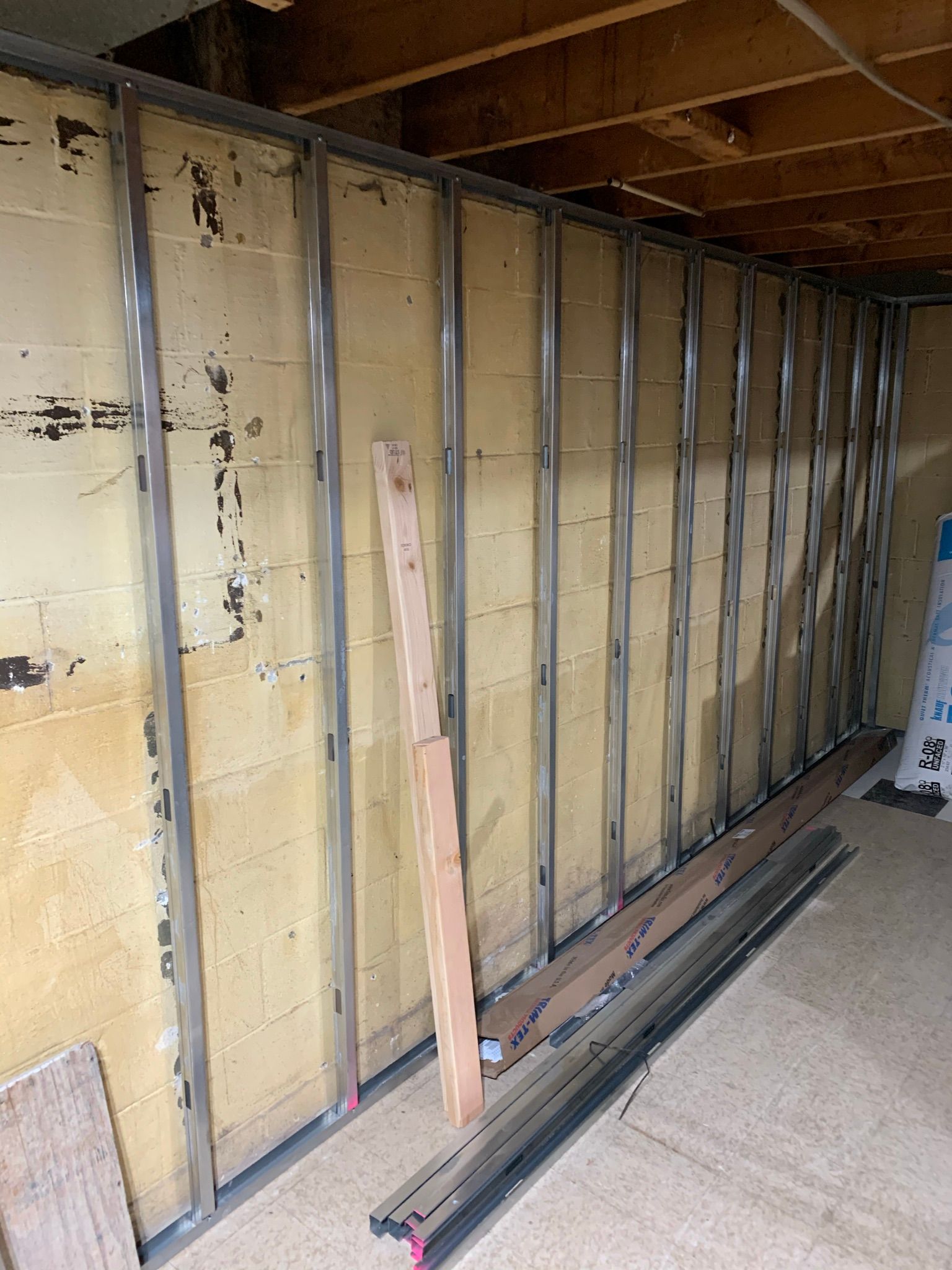Metal studs frame a basement wall. Beige cinder block wall with wood beams above.