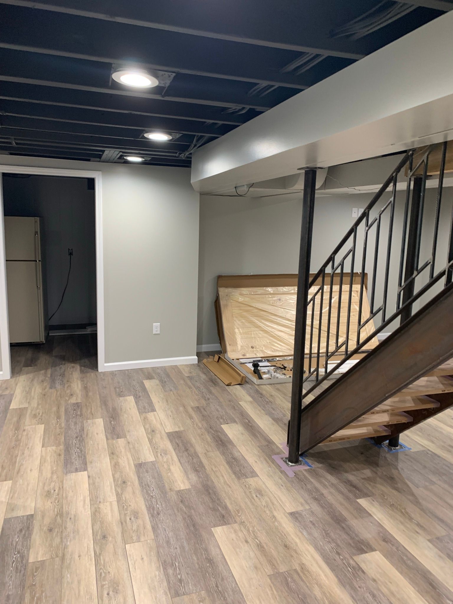 Basement with wood-look floor, stairs with metal railing, and doorway to another room with refrigerator.