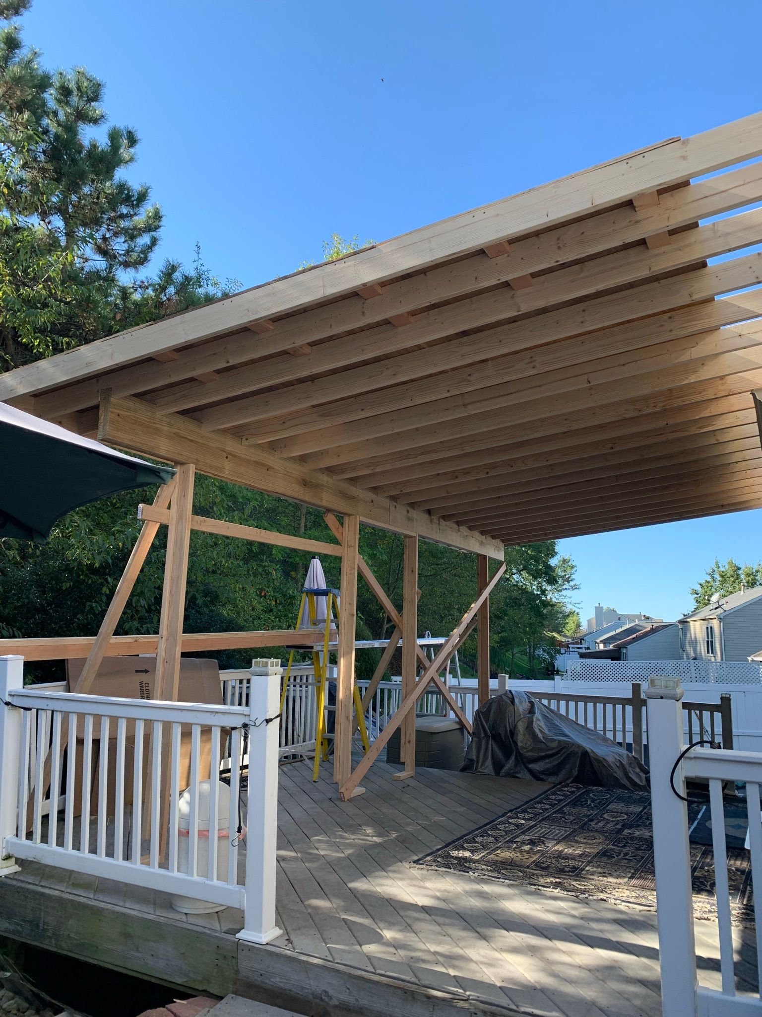 A wooden pergola being built on a deck, supported by temporary braces. Bright blue sky overhead.