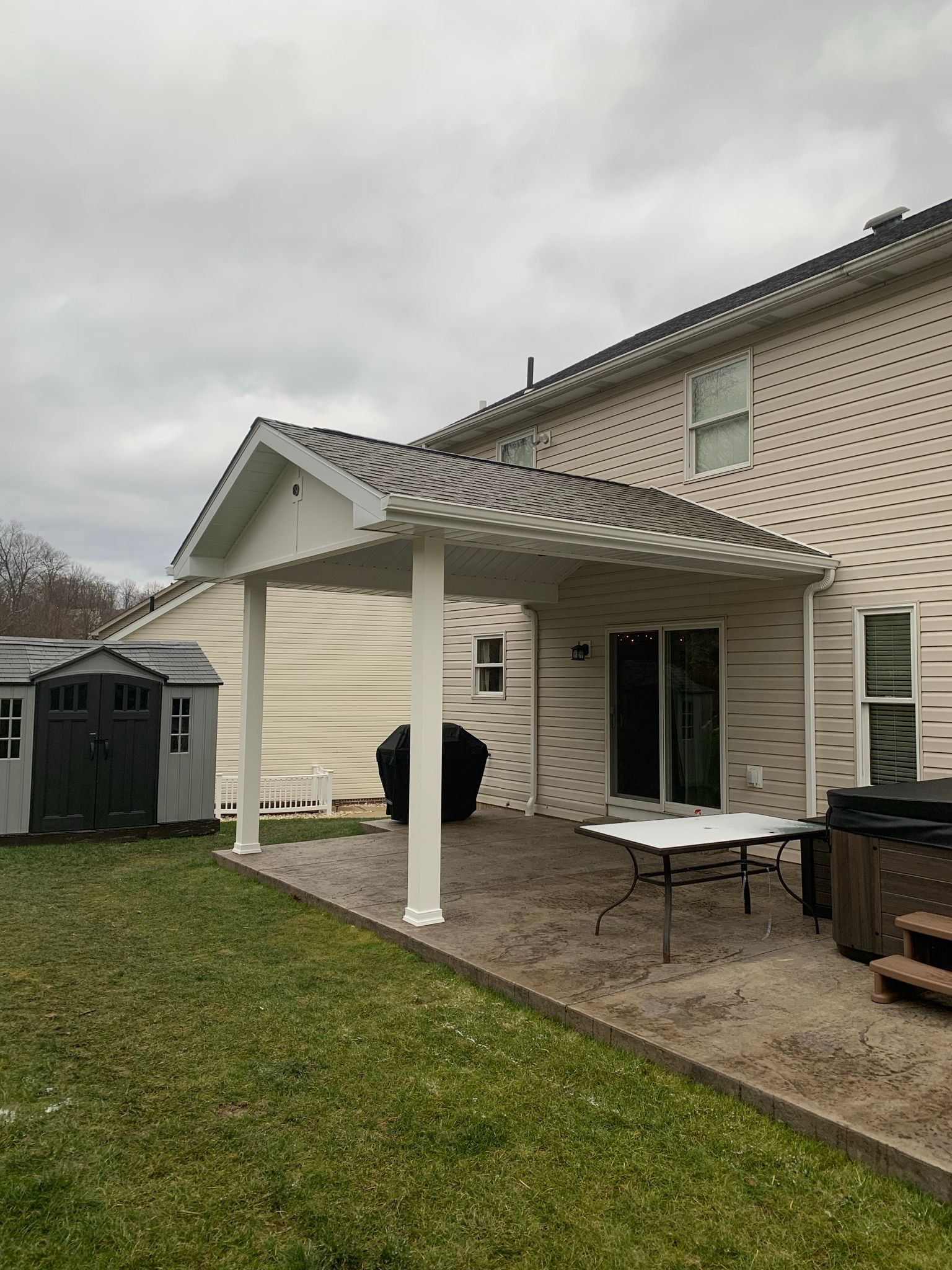 White patio cover attached to a two-story house. Gray paver patio with table and hot tub, green lawn, and shed.