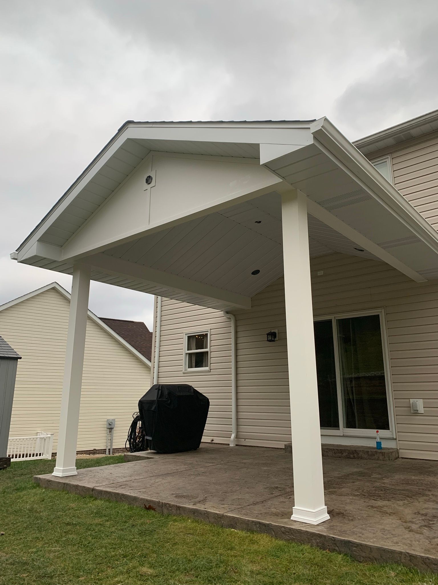 White covered patio attached to a house with a grill on a brick patio.
