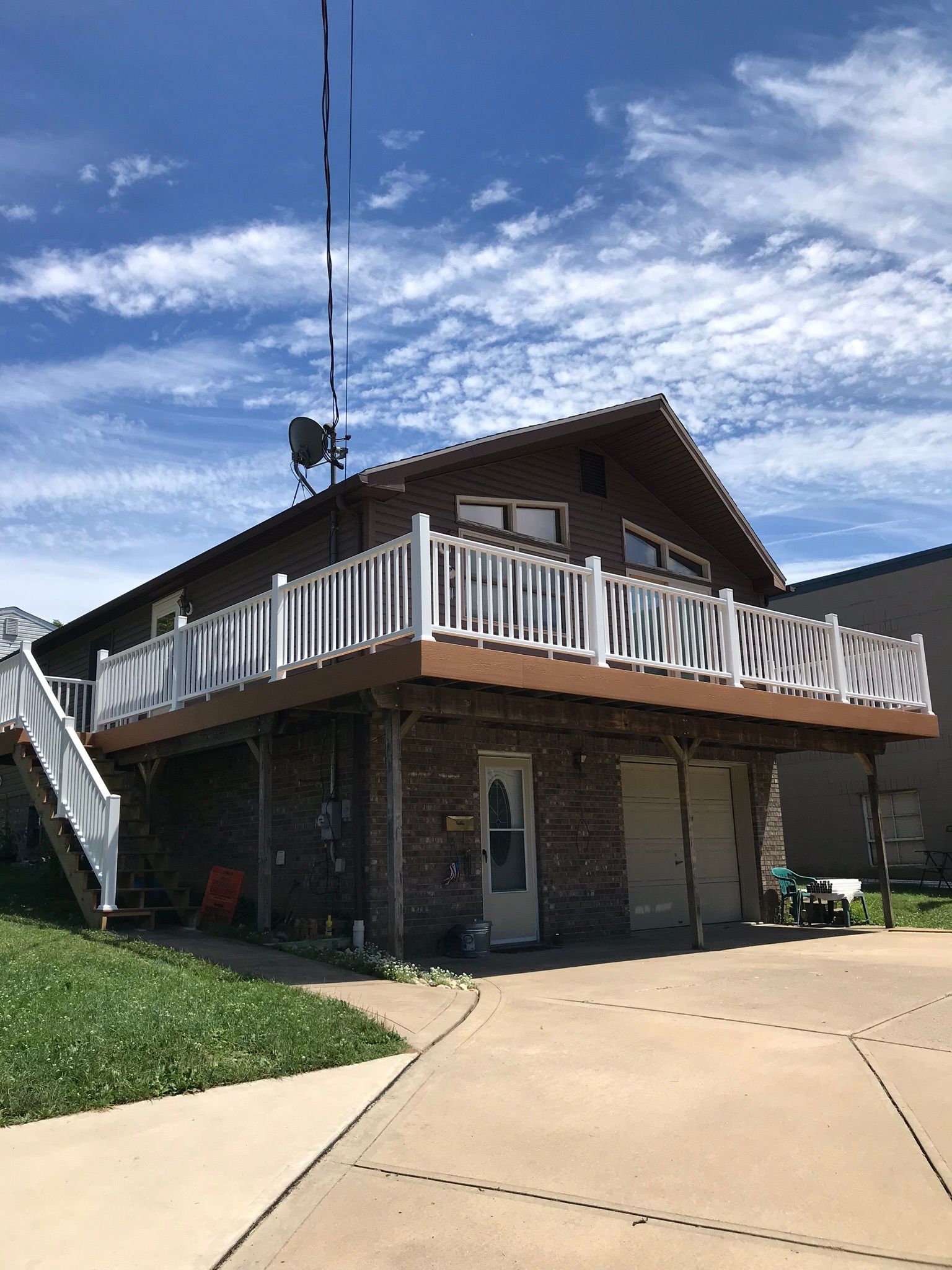 Two-story house with a deck and white railings.  Brick base, brown siding, and a driveway. Blue sky with clouds.