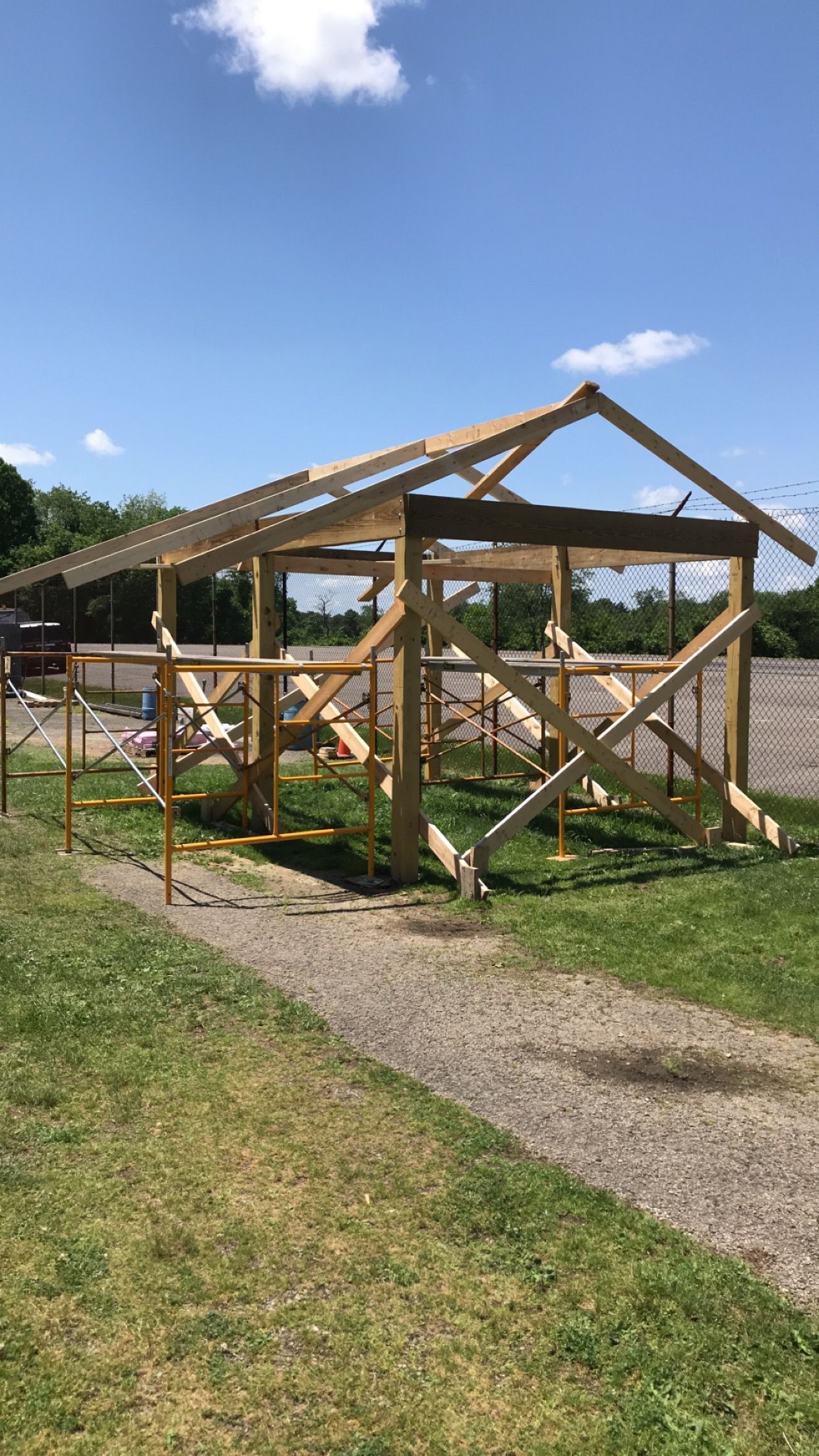 Wooden structure under construction outdoors, on gravel path and grass, against a blue sky.