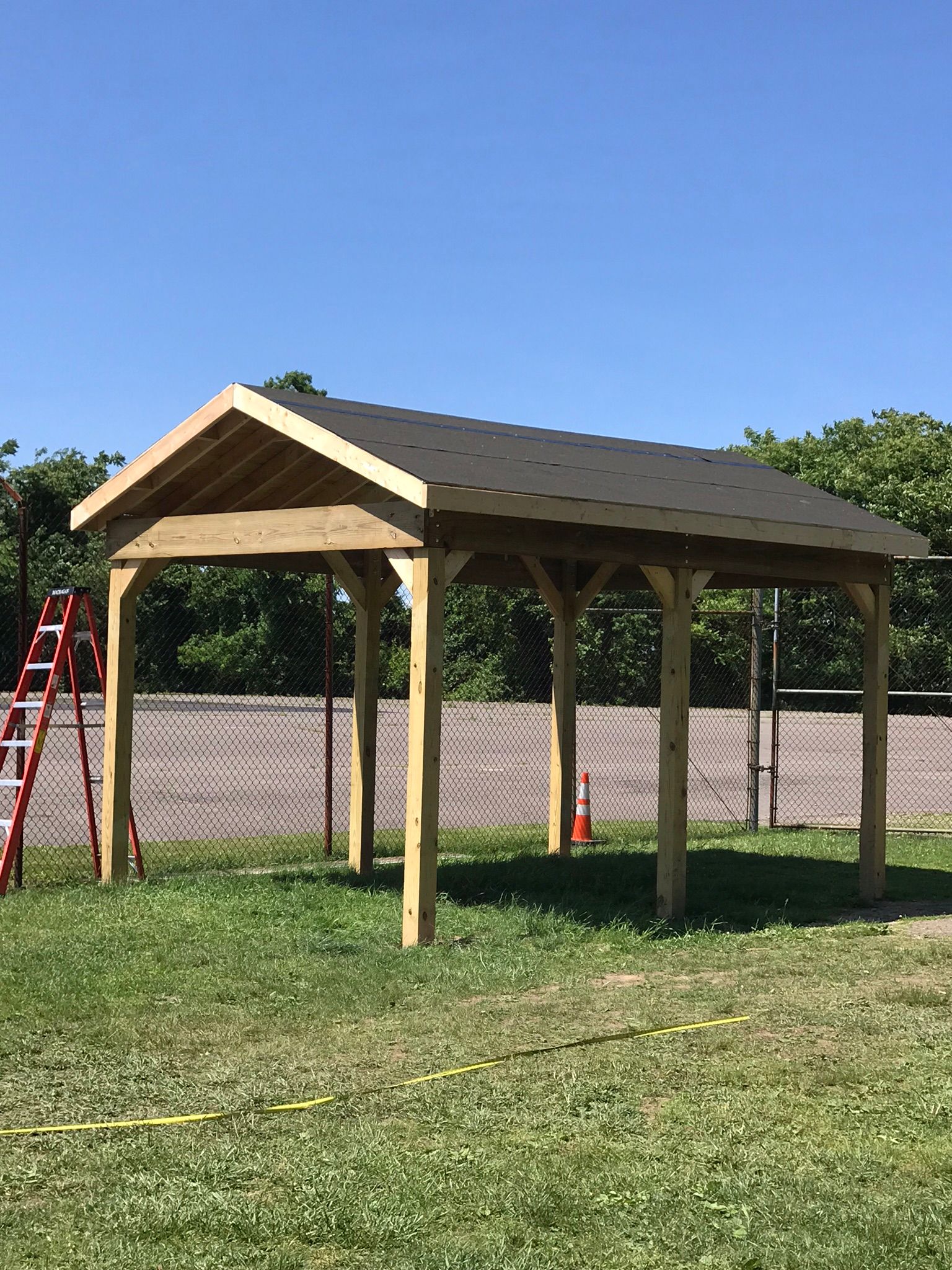 A wooden gazebo with a black roof stands on grass, next to a chain-link fence and a ladder.