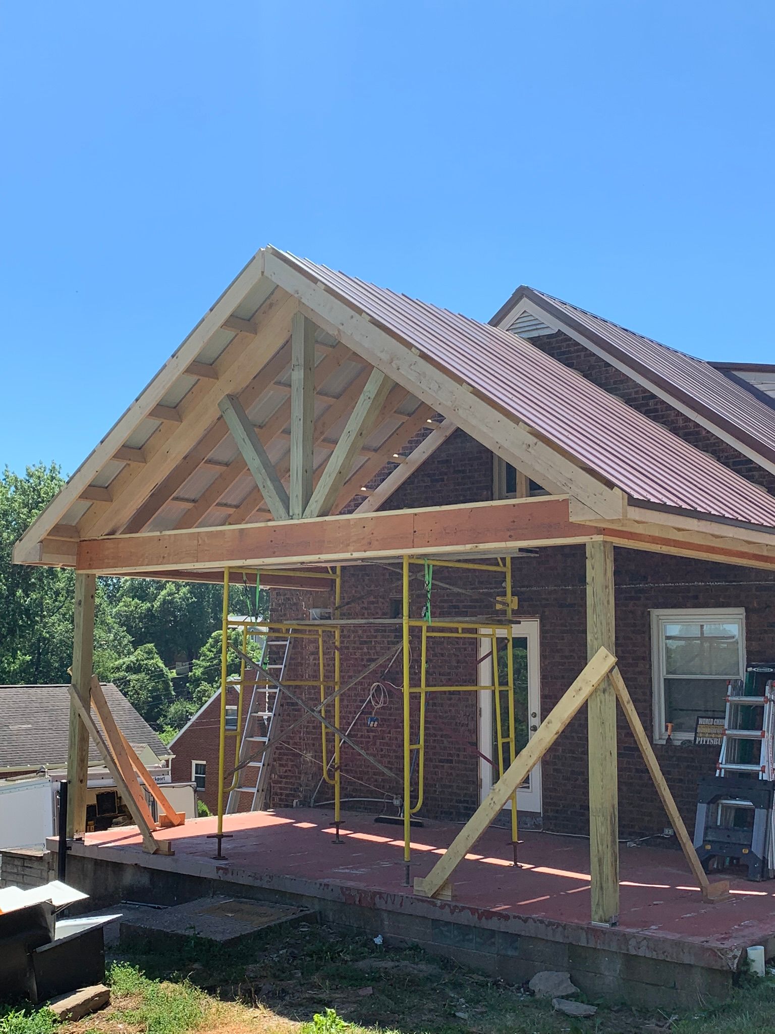 Construction of a porch roof, with exposed wooden beams and trusses, in front of a brick building.