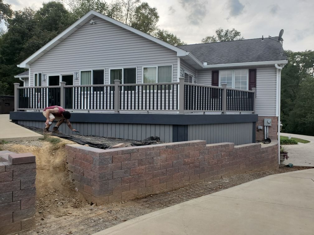Person working on a brick retaining wall in front of a house with a deck; outdoor setting.