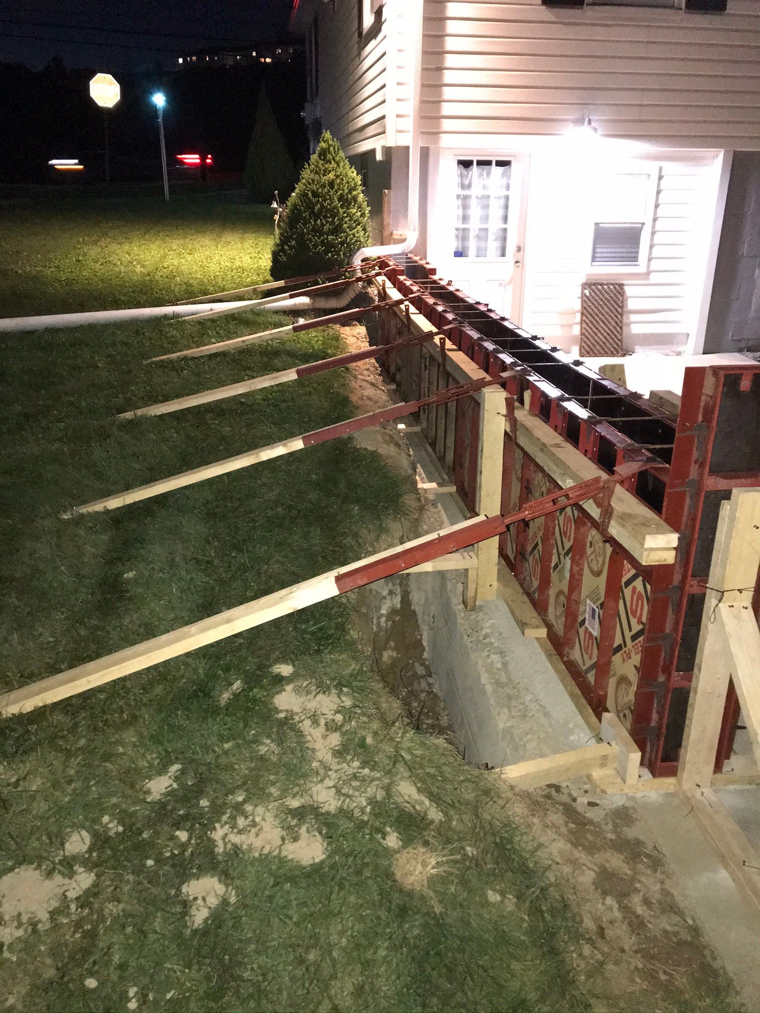Wooden forms supported by braces sit in front of a building foundation, construction at night.