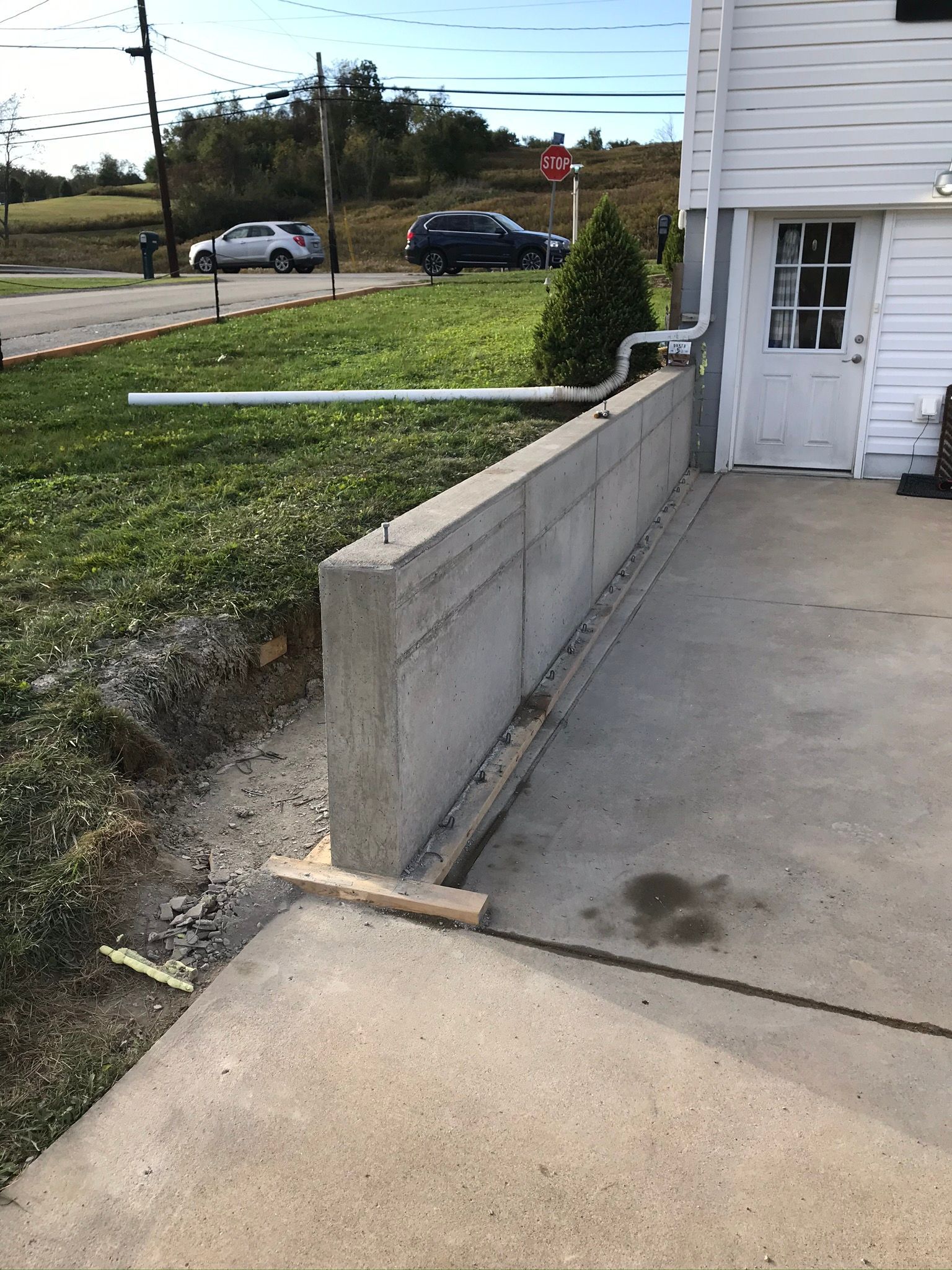 Concrete retaining wall next to a driveway, grass, and a white building with a door; cars in background.