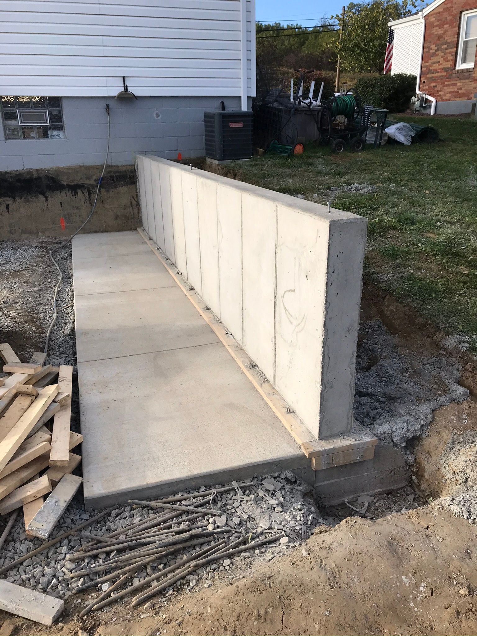 Concrete retaining wall under construction next to a house. Rebar, lumber, and gravel are visible.