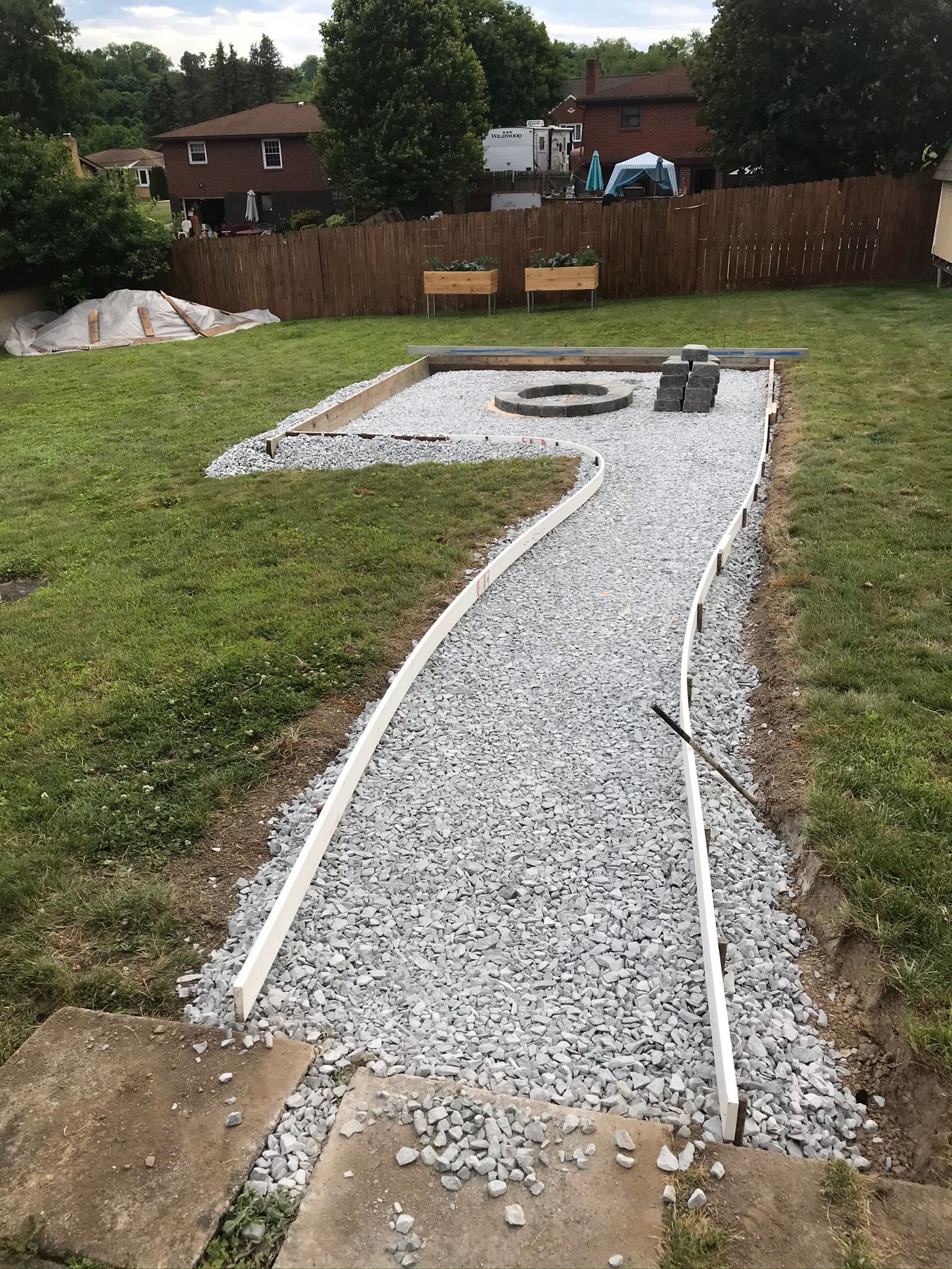 A gravel path leads to a fire pit in a backyard, bordered by white edging and surrounded by grass.