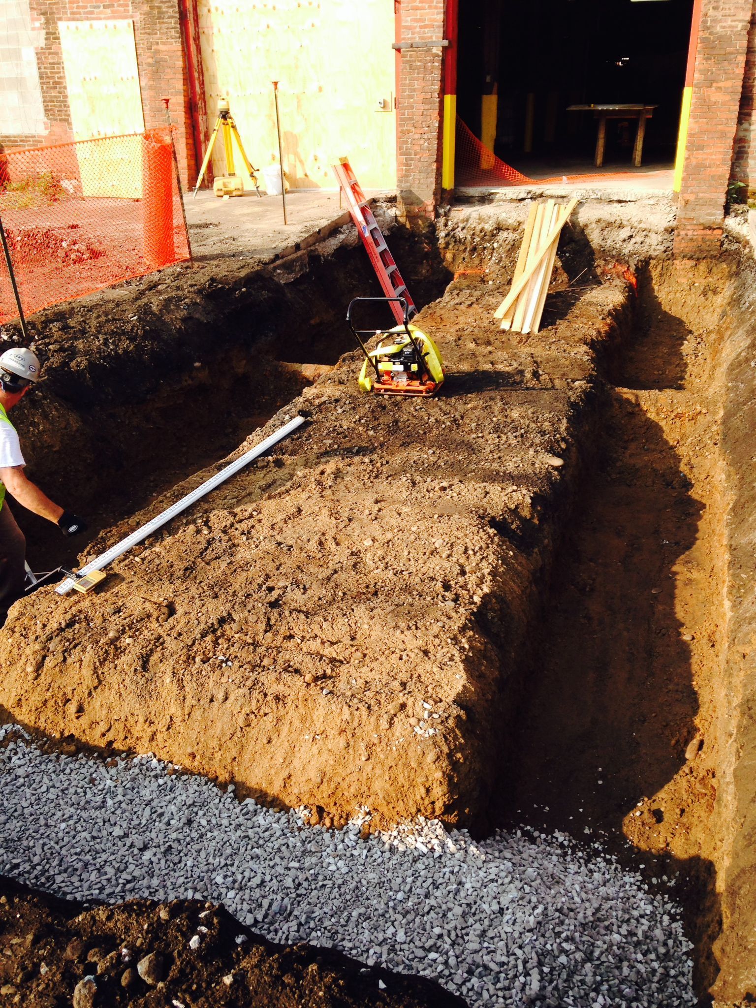 Construction site with trenches, worker, machinery, and building entrance.