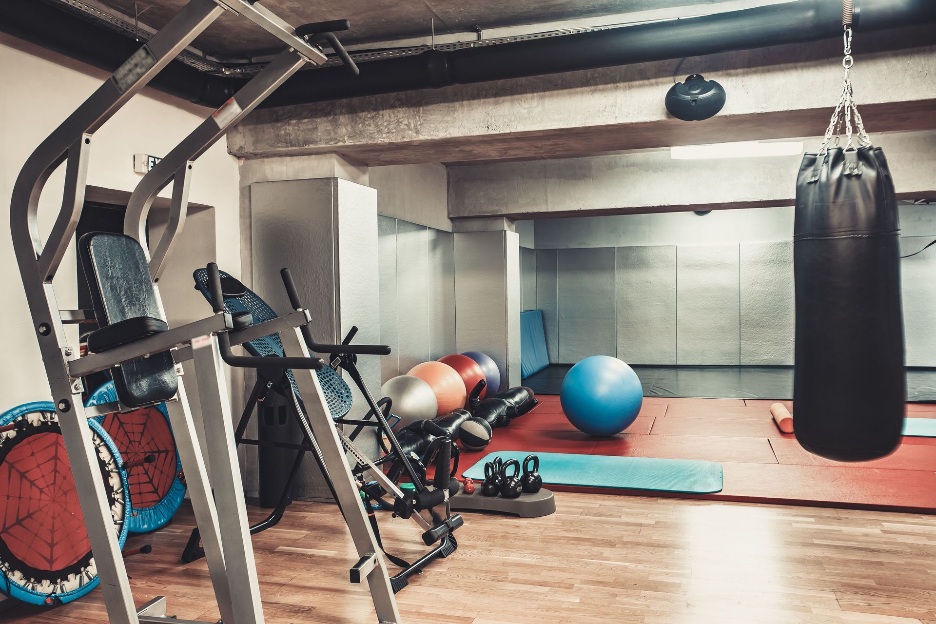Gym with pull-up bars, punching bag, exercise balls, and mats on wooden floor.