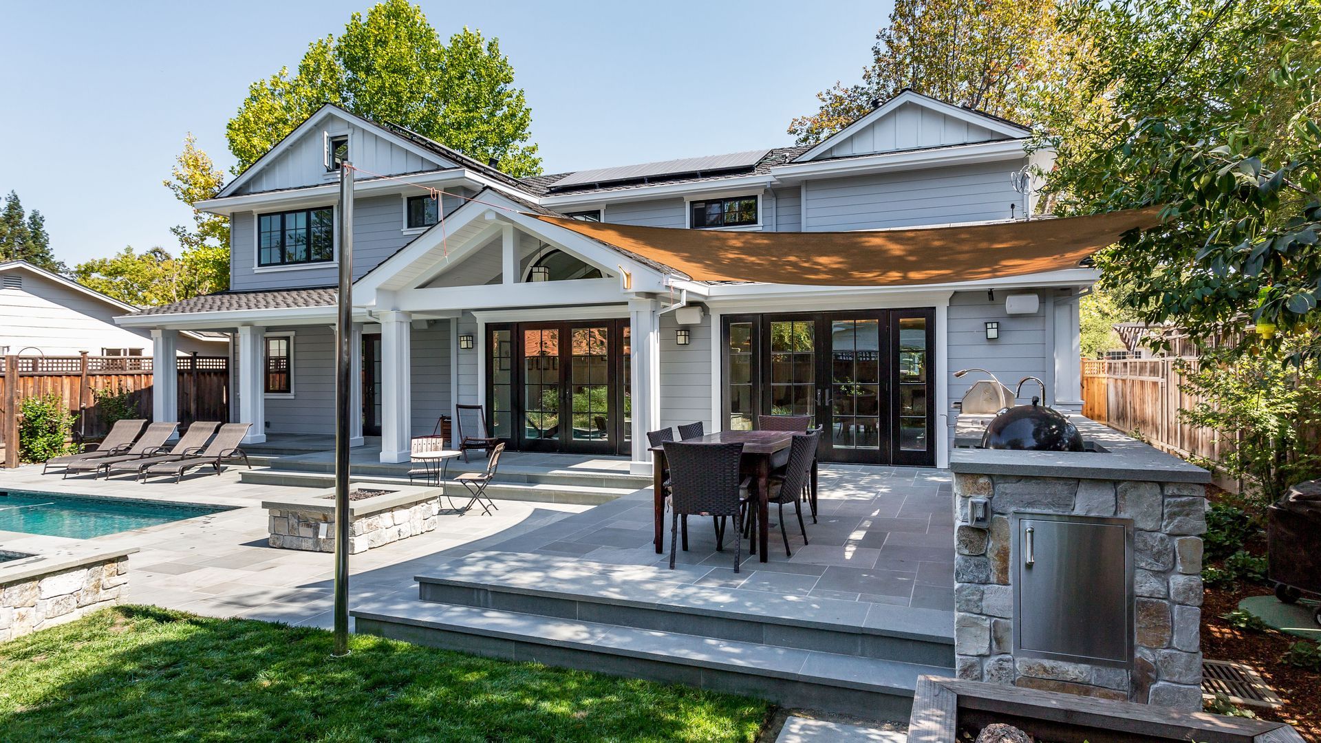 Backyard with a pool, patio, outdoor kitchen, and gray house with black-framed windows.