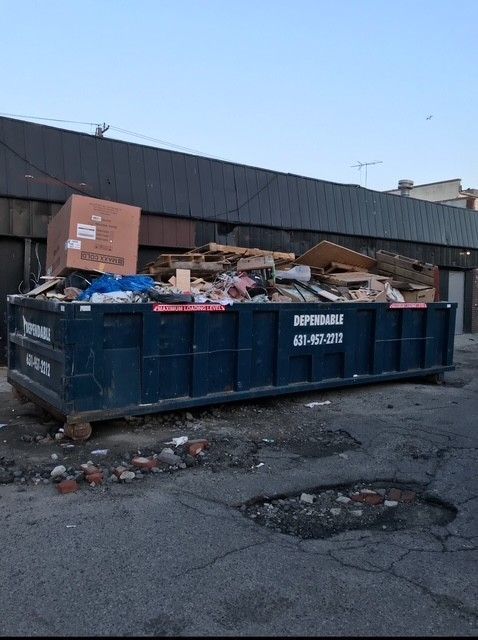 A full, dark blue dumpster on a cracked street, filled with debris. A large box sits on top.