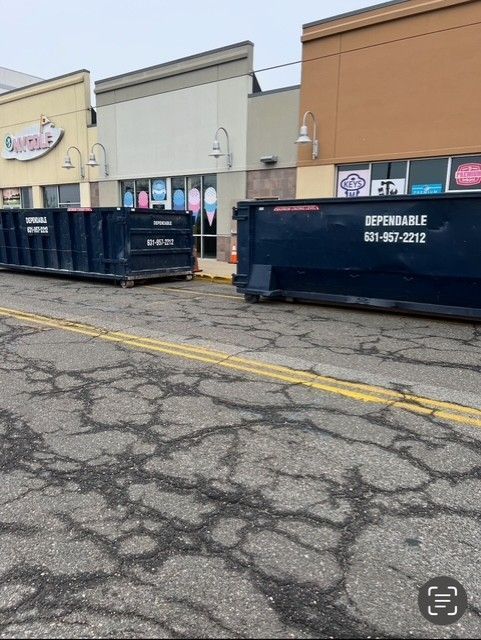Two blue dumpsters parked in front of storefronts on a cracked asphalt street.