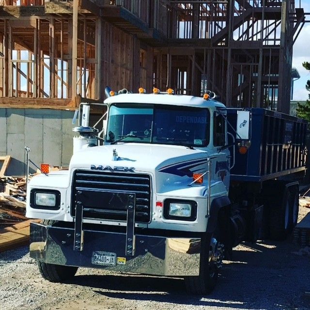 White Mack dump truck parked in front of a partially built wooden house. Blue and silver accents on truck.