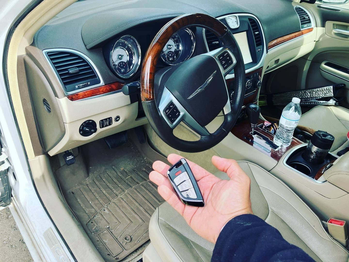 Car interior with steering wheel and dashboard; a hand holds a car key fob near the driver’s seat.