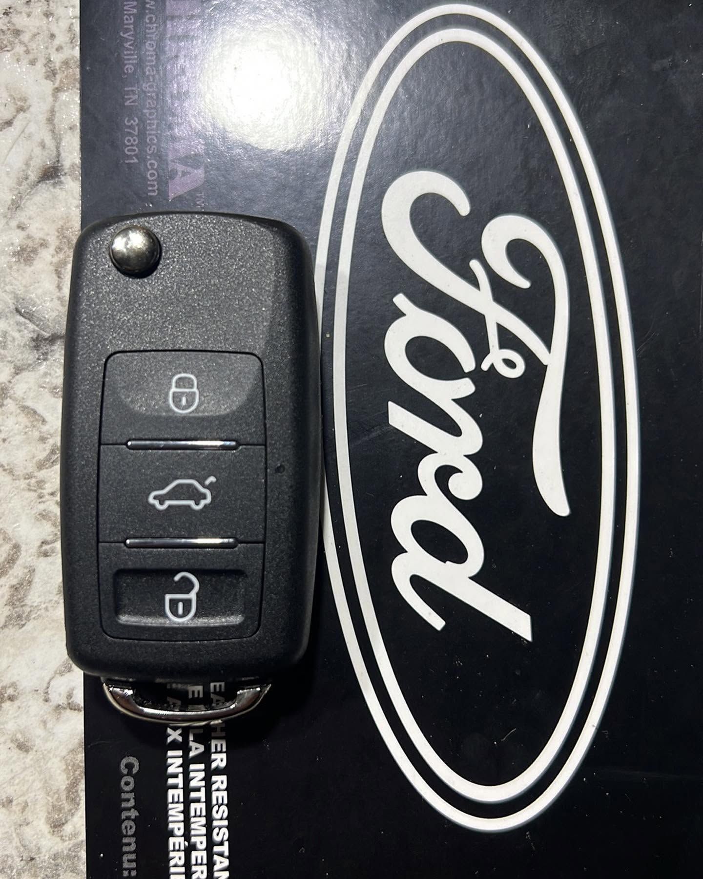 Ford car key fob on a black Ford-branded background, showing lock, trunk, and unlock buttons.