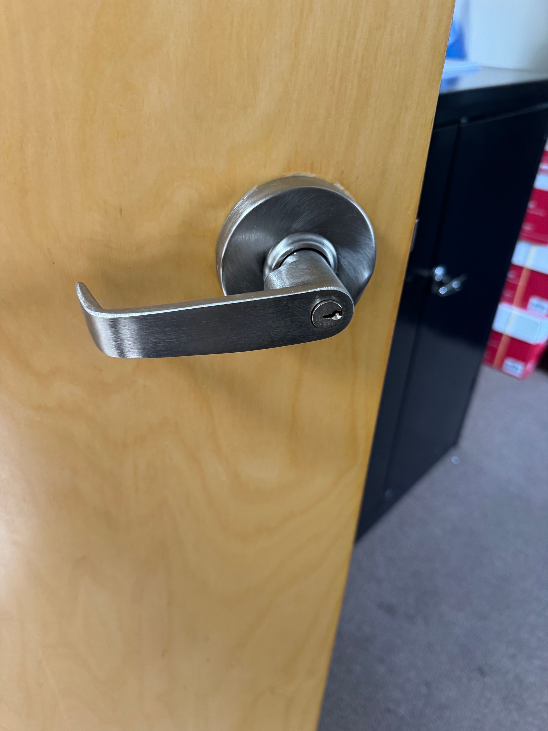 Close-up of a silver door lever on a light wood door, with a red-and-blue object in the background