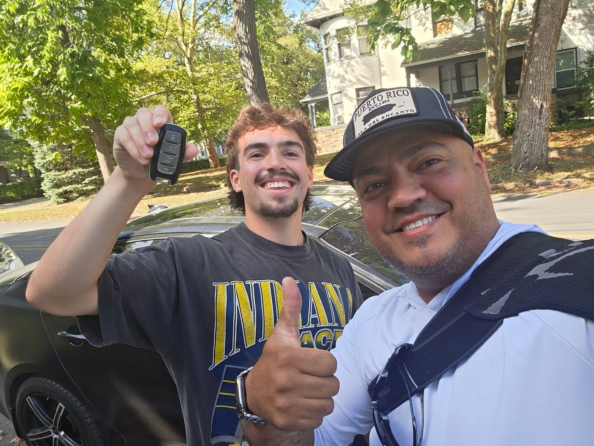 Two smiling people outdoors, one holding car keys and the other giving a thumbs-up.