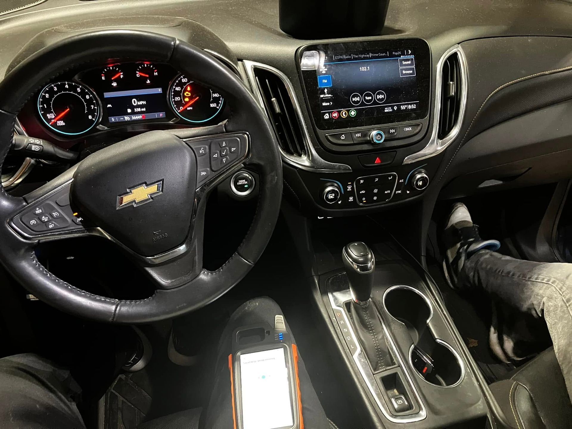 Inside a Chevrolet car, dashboard and steering wheel visible with a person’s legs in the driver’s seat, phone on lap