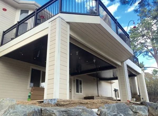 A two-story house with a deck and black railing. The underside of the deck is dark, supported by white columns.