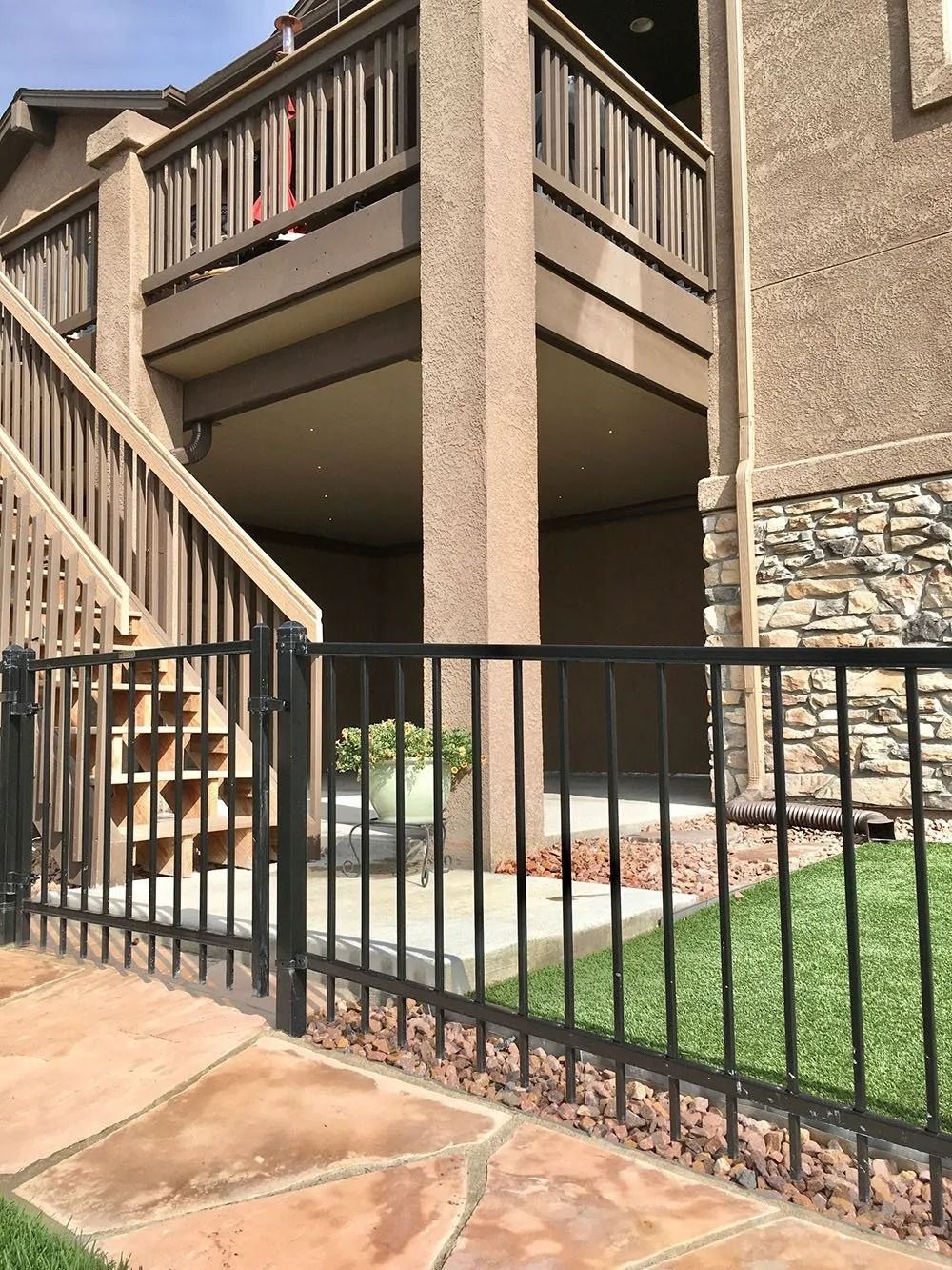 Black metal fence in front of a building with a deck and stairs. The building exterior is tan with some stone.