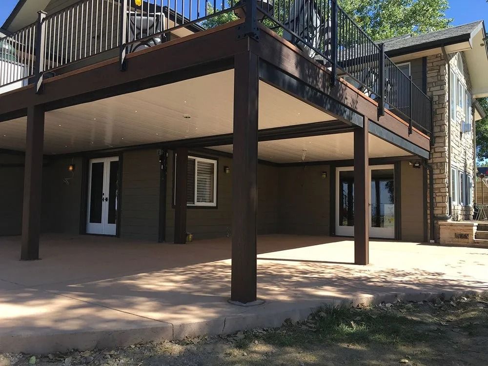 A house with a large, dark-brown deck supported by pillars. The underside of the deck forms a sheltered patio area.
