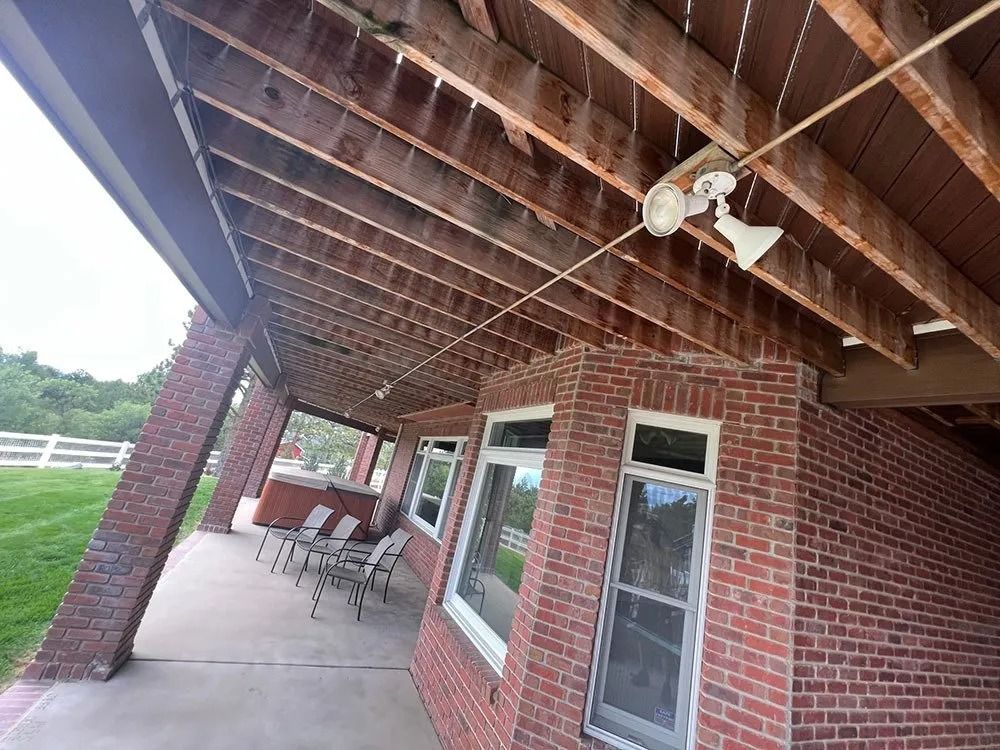 Brick building with an overhanging wooden roof, a covered porch, and windows. A grassy area and white fence are in the background.