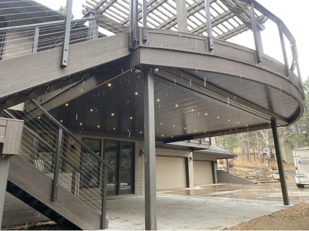A multi-level deck with a curved design, cable railings, and string lights. It is attached to a house with a garage, viewed outdoors.