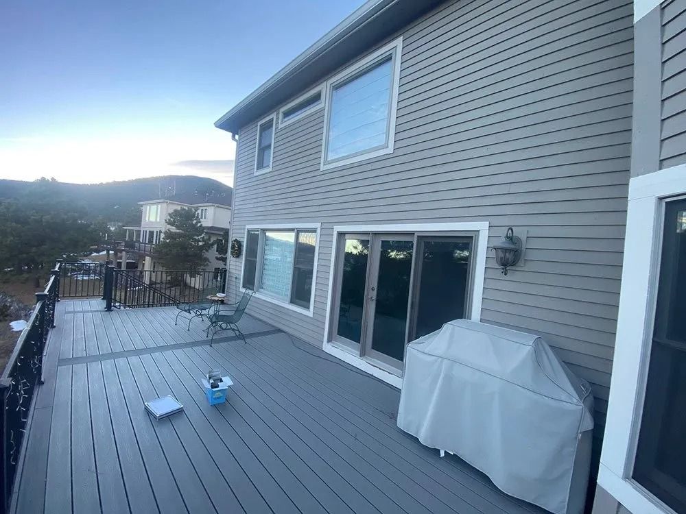 A two-story house with a gray deck, sliding glass doors, and a covered grill. In the background are trees and mountains.