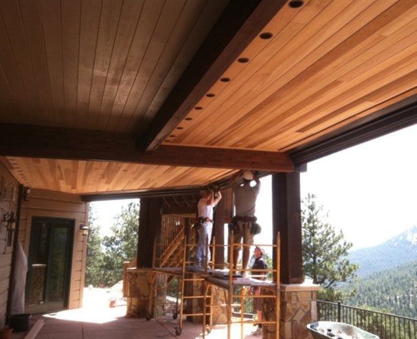 Construction workers installing a wooden ceiling on a covered patio with a scenic mountain view. One worker stands on a scaffold, while another works from ground level.