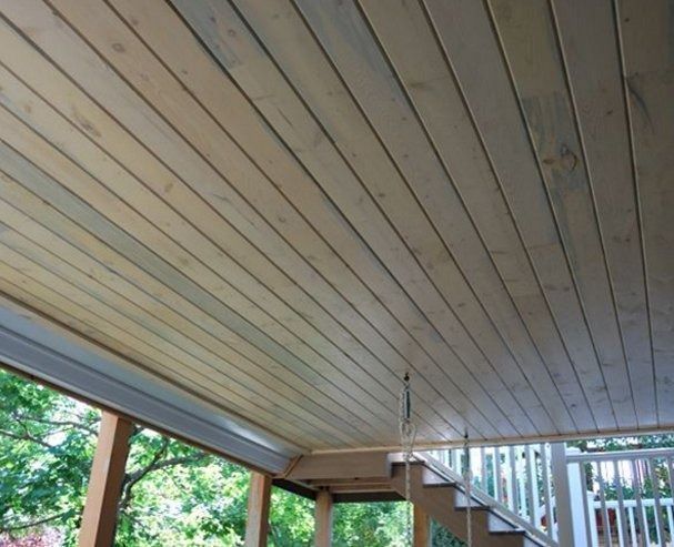 Wooden deck ceiling with gray-toned planks. Light shines from the open side, near a staircase and trees.