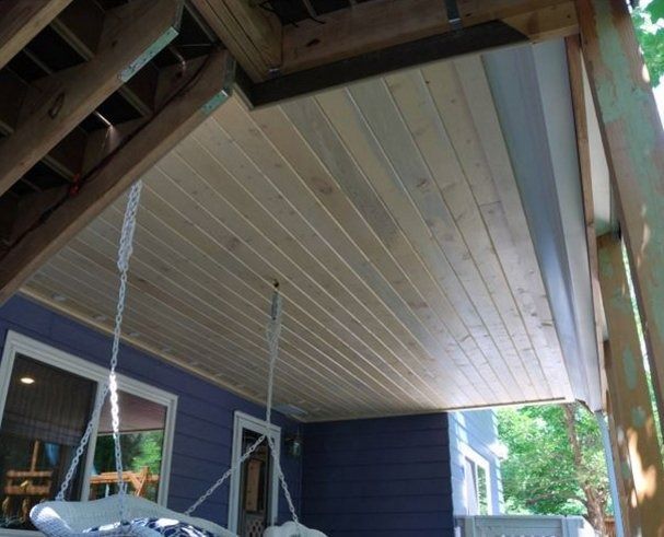 A wooden porch ceiling with light-colored planks. A porch swing hangs below the ceiling; a blue wall with windows is visible.