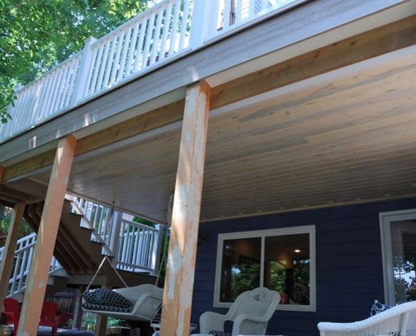 Wooden deck with white railing, supported by wooden posts, overlooking a blue house with windows and chairs.