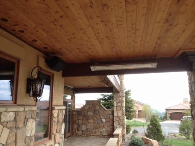 An outdoor porch with a wooden ceiling, stone walls, and a view of the surrounding landscape. There is a light fixture and a mounted speaker.