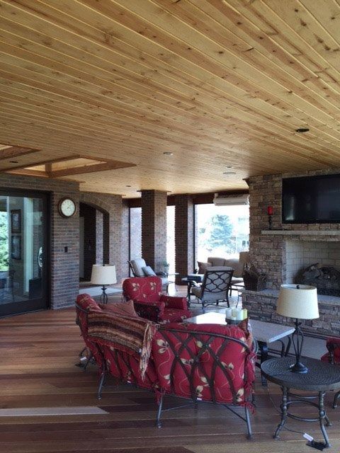 An outdoor living room with a wood ceiling and floor, red patterned chairs, a stone fireplace, and large windows.