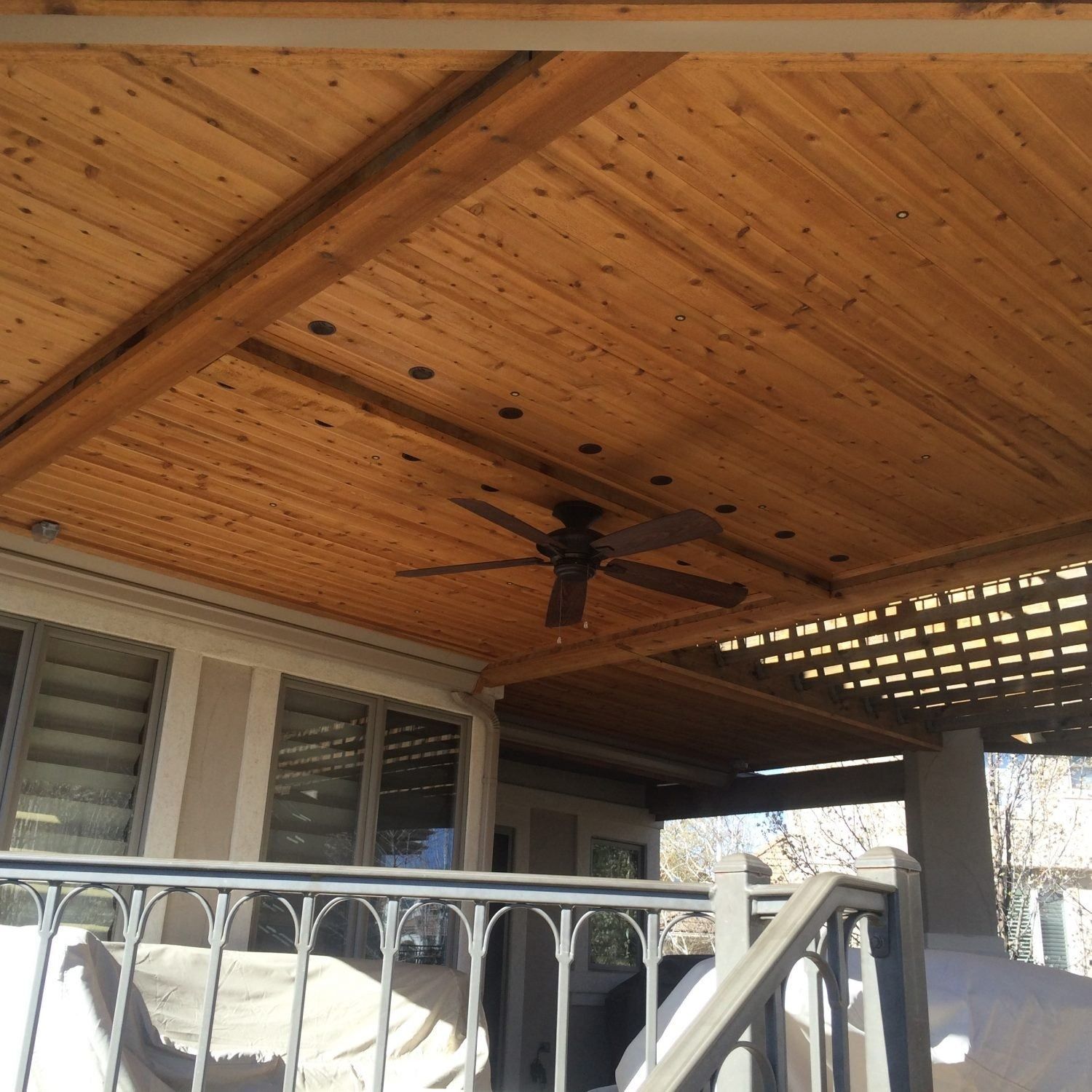 A wooden patio ceiling with a ceiling fan and a latticed section, viewed from below. The balcony has a black wrought-iron railing.
