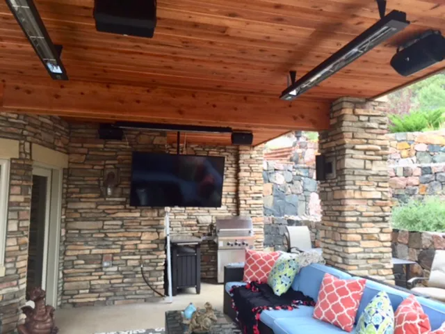 Outdoor patio with TV, speakers, heaters, and stone walls. A blue sofa has colorful throw pillows.