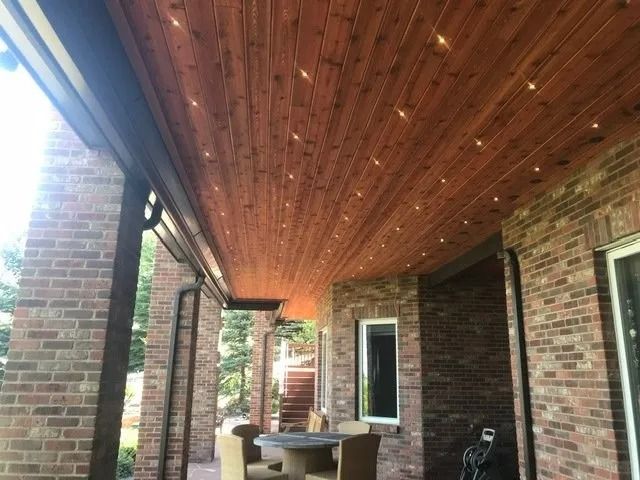 A brick patio ceiling with wood paneling is adorned with string lights. The patio is partially covered and has brick columns.