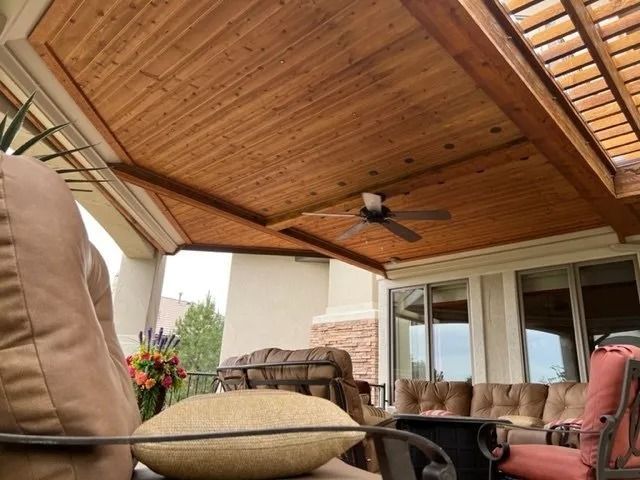 A patio with a wooden ceiling, a ceiling fan, and outdoor seating. A sunny day is visible through glass doors.